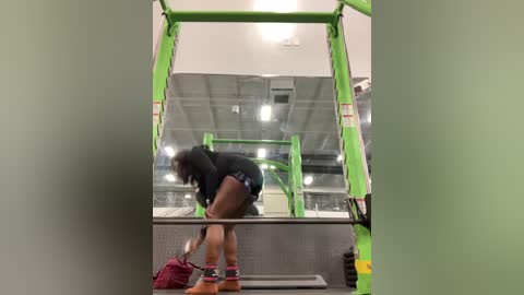 Media: Video of a muscular woman lifting weights in a gym with a green lifting rack. She wears a black hoodie and shorts, and is holding a maroon backpack.