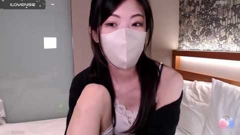 Media: Video of an East Asian woman with long black hair, wearing a white face mask, black top, and lace bra, in a modern bedroom with white bed and grey wall art.