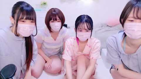 Media: Video of four young Asian women in casual, light-colored clothing, wearing masks, sitting closely together, indoors, with a blurred background.