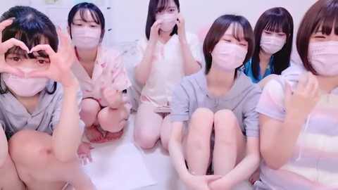 Media: Video of six young Japanese women in surgical masks, casual clothes, seated on a white floor, making heart shapes with their hands, creating a sense of unity and solidarity.