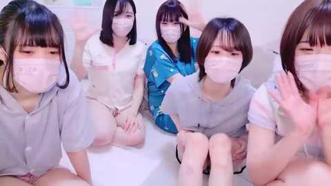 Media: Video of five Asian women, all wearing surgical masks, seated on a white surface, dressed in various casual outfits. They have different skin tones and facial features, suggesting a diverse group.