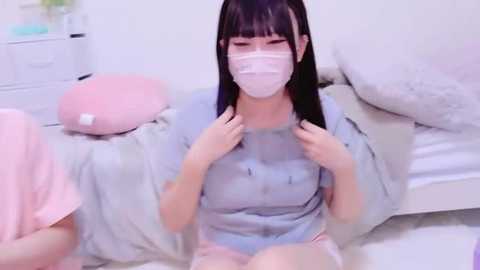 Media: Video of a young East Asian woman with straight black hair and a white surgical mask, wearing a light blue shirt, seated on a fluffy white rug in a pastel-colored bedroom.