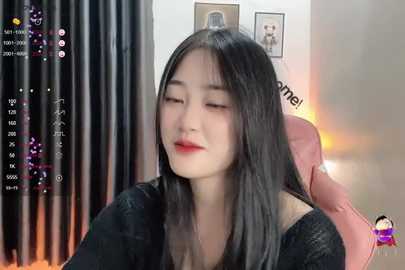 Media: A video of an East Asian woman with long black hair, wearing red lipstick, seated on a pink gaming chair, surrounded by dimly lit room with black curtains and framed pictures on the wall.