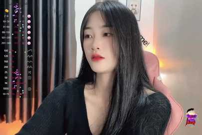 Media: Video of an East Asian woman with long black hair, fair skin, and red lipstick, wearing a black top, sitting on a pink chair, in a dimly lit room with black curtains.