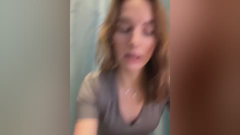 Media: A blurred video of a woman with light skin and shoulder-length brown hair, wearing a grey top, taken from a close-up angle, possibly in a bathroom.