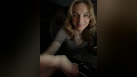 Media: Video of a blonde woman with wavy hair, wearing a dark gray top, sitting in a dimly lit car, smiling slightly, hand raised.