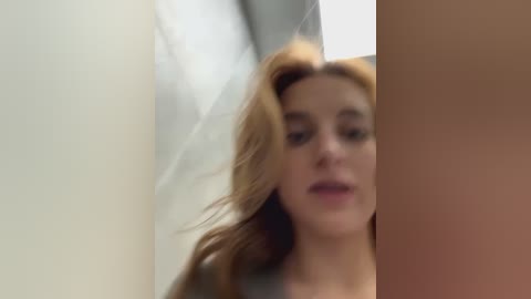 Media: A blurry video of a woman with long, wavy, light brown hair, looking down. She is partially visible behind a vertical object. The background is an indoor, minimalist setting with light-colored walls.