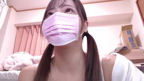 Media: Video of a young Asian woman with long brown hair in pigtails, wearing a pink face mask, white tank top, and white jacket, in a cluttered bedroom with pink curtains, cardboard box, and a bed with white sheets.