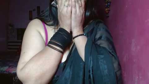 Media: Video of a woman with medium skin tone, dark hair, wearing a black robe and pink bra, covering her face with hands, standing against a pink wall, bracelets on wrists.