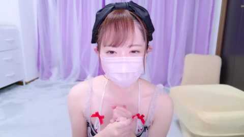 Media: Video of an Asian woman in a maid outfit, wearing a white face mask, black headband, and apron with red bows. She kneels on light gray flooring. Background includes white dresser and purple curtains.