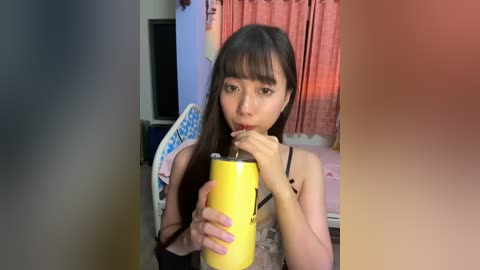 Media: Video of a young Asian woman with long dark hair, wearing a sleeveless top, drinking from a yellow cup, sitting in a room with a pink curtain and a TV in the background.