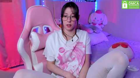 Media: A video of an East Asian woman with glasses and a white t-shirt featuring a pink character, seated in a pink gaming chair.