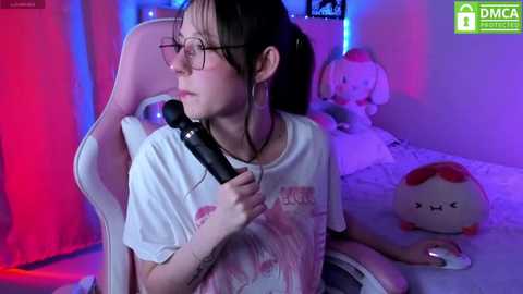 Media: A video of a young woman with glasses, dark hair, and a white T-shirt, sitting in a pink gaming chair, holding a microphone, surrounded by plush toys in a brightly lit, colorful bedroom.