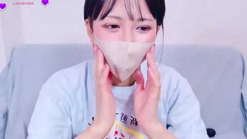 Video of an Asian woman with fair skin, wearing a light blue shirt and beige face mask, holding her cheeks. Background is a light-colored sofa.