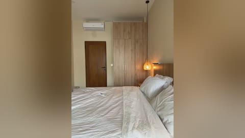 Media: Video of a minimalist, modern bedroom with beige walls, white linens, wooden wardrobe, bedside lamp, and air conditioner.