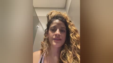 Media: Video of a young woman with curly, wavy, light brown hair and a medium complexion, wearing a light-colored top, standing in a dimly lit hallway with beige walls and a recessed ceiling light.