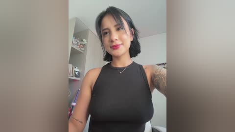 Media: Video of a Latina woman with medium skin tone, short black hair, wearing a black sleeveless top, standing in a minimalist room with white walls, shelves, and a mirror.