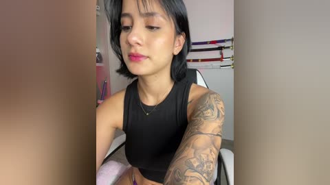 Media: Video of a young woman with short black hair, wearing a black tank top, revealing intricate tattoo sleeves on her left arm. She has a light complexion, wearing subtle makeup and a relaxed expression. The background shows a simple room with a white wall and a few items.