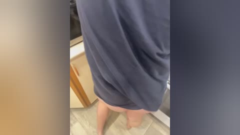 Media: Video of a person standing in a bathroom, wearing a dark blue shirt, no pants, bare feet on beige tiles, wooden cabinets and white sink visible in the background.