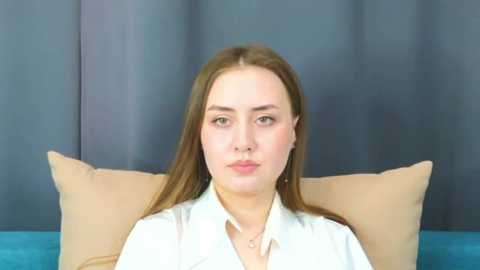 Media: Video of a Caucasian woman with fair skin and straight, light brown hair, wearing a white blouse, seated against a blue curtain backdrop.