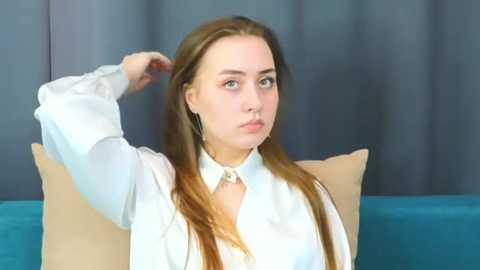 Media: Video of a young Caucasian woman with long brown hair, wearing a white blouse, sitting on a blue couch against gray curtains. She looks contemplative.