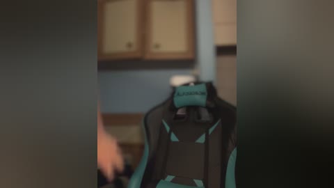 Media: Video of a black gaming chair with teal accents and the brand \"Logitech\" in a dimly lit kitchen. The background features beige wooden cabinets and a blurred stove. The image has a slight blur effect.