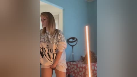 Media: Video of a blonde woman in a glittery sweater, partially undressed, standing in a dimly lit bedroom with a red polka-dotted bedspread.