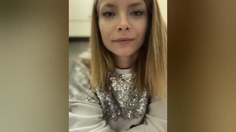 Media: A video of a fair-skinned, blonde woman with a serious expression, wearing a sequined, silver top, standing indoors against a blurred brown and beige background.