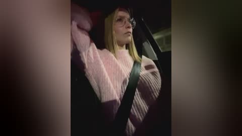 Media: Video of a blonde woman in a pink knit sweater and black headscarf, driving a car with a black seatbelt visible, dark interior, and blurred background.