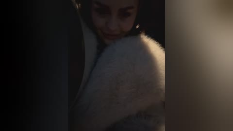 Media: A dimly lit video shows a close-up of a woman with light skin and dark hair, partially obscured by a fluffy white fur collar, creating a dramatic, intimate atmosphere.