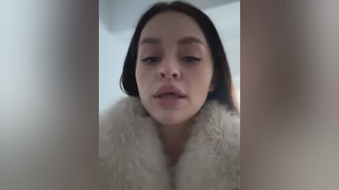 Media: Video of a young woman with fair skin and straight brown hair, wearing a white fluffy coat, seen from a close-up angle, slightly blurred.