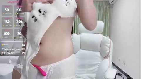 Media: A video of a light-skinned woman lifting a white shirt with cat ears, revealing her midriff and pink underwear. She stands in a modern, white-walled room with a white leather armchair and light green curtains.