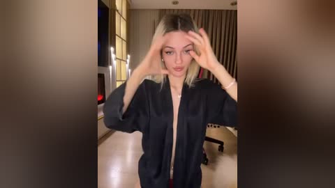 Media: A video of a young woman with straight, shoulder-length blonde hair, wearing a black satin robe, adjusting her hair in a modern, warmly-lit living room.