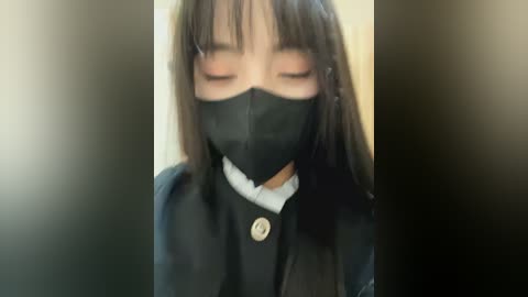 Media: Video of an Asian woman with long, straight black hair, wearing a black face mask, black cardigan, and white collared shirt. She appears to be standing in an indoor setting with blurred background.