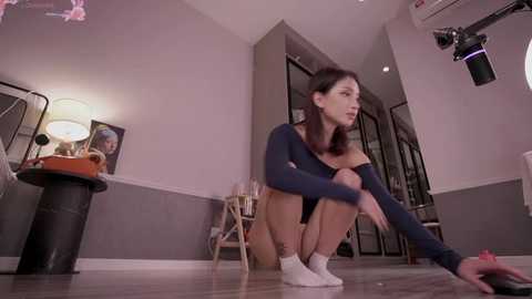 Media: Video of a woman with light skin, brown hair, and slender physique, crouching on a wooden floor, wearing a dark blue long-sleeve top and white socks. She is tying her shoelace. Background features a modern room with gray walls, a white lamp, and a partially visible mirror.