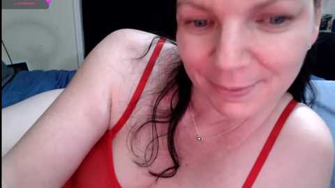 Media: Video of a fair-skinned woman with shoulder-length black hair, smiling slightly, wearing a red tank top, lying on a bed with blue sheets, and a black door in the background.