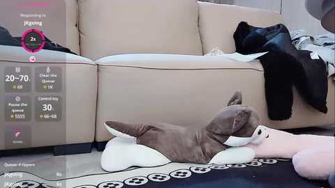 Media: Video of a brown and white plush dog toy curled up on a black and white patterned rug in a modern living room with beige couch and scattered clothing.