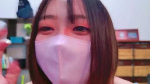 Media: Video of an East Asian woman with shoulder-length dark hair, wearing a white face mask, and pink eyeshadow. Background features a cluttered room with shelves, books, and boxes.