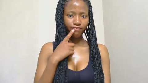 Media: Video of a young Black woman with braided hair, medium skin tone, wearing a dark sleeveless top, covering her chest. She holds her index finger to her lips in a thinking pose against a plain beige wall.