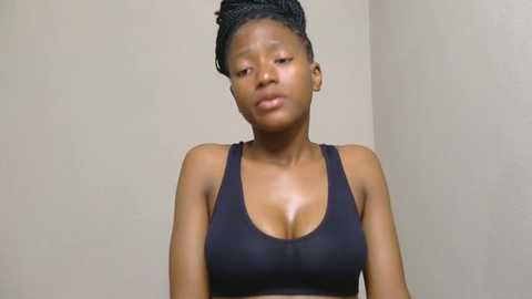 Media: Video of a young Black woman with medium-dark skin tone, wearing a black sports bra, standing against a plain beige wall, her hair styled in a neat bun.