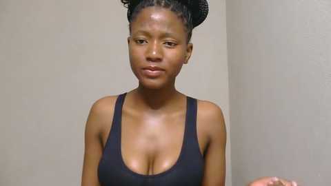 Video of a dark-skinned woman with braided hair, wearing a black tank top, standing against a plain beige wall. Her expression is neutral, and she appears to be holding something in her hand.