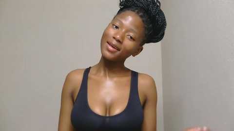 Media: Video of a dark-skinned woman with braided hair, wearing a black tank top, standing against a plain beige wall. She has a serene expression, with a slight smile.