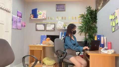 Media: A video of an Asian woman with a mask and long black hair, seated at a desk in a cluttered office, surrounded by paperwork, plants, and a computer.