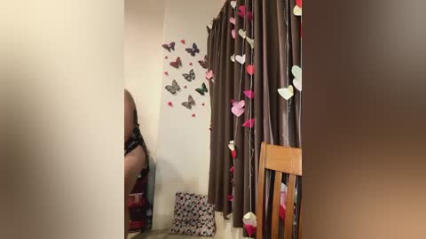 Media: A video of a bedroom corner with brown curtains adorned with pink and white butterflies, a small wooden chair, and a partially visible person wearing black lingerie.