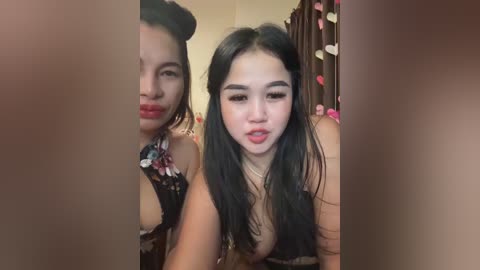 Media: Video of two Asian women with long black hair, one wearing a floral dress, the other a black top. They have light skin and are smiling. Background includes a heart-patterned curtain.