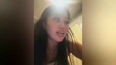 Media: A video of a young woman with long black hair, wearing a white top, leaning forward with a slightly open mouth, taken in a dimly lit room with beige walls and a ceiling light.