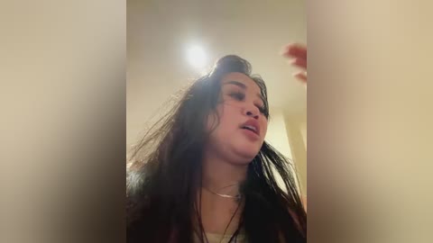 Media: Video of a young woman with long, dark hair, slightly messy, and a neutral expression, wearing a white top and necklace, captured from a low angle, indoors.