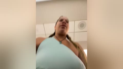 Media: A low-angle video of a middle-aged woman with light skin and auburn hair, wearing a light blue top, standing in a beige bathroom with white tiles and a circular ceiling vent.