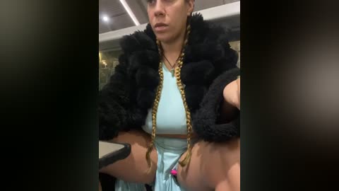 Media: Video of a woman with short brown hair, wearing a light blue top, black faux fur coat, and long braided hair with gold chains, sitting on a bus.