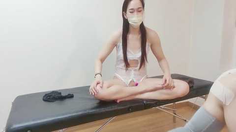 Media: Video of an Asian woman with long black hair, wearing a white lace bra and panties, seated cross-legged on a massage table, applying lotion to her legs, with a mask and bracelet.